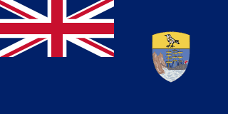 The flag of Saint Helena, Ascension and Tristan da Cunha is blue with the UK flag in the canton and the national coat of arms shield centered on the fly half. The upper third of the shield depicts a white plover, a native bird, on a yellow field. The rest of the shield depicts a rocky coastline and a three-masted sailing ship with sails furled and flying an English flag.
