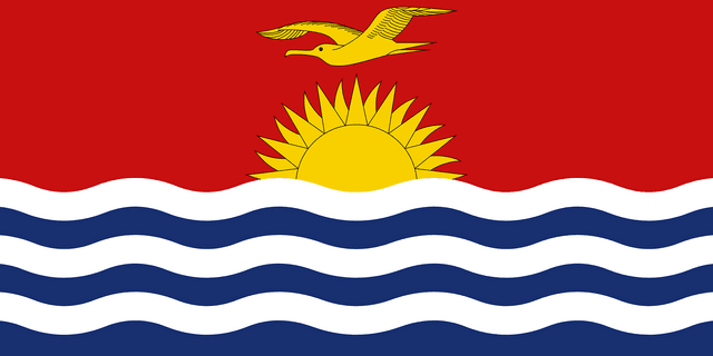 The flag of Kiribati is divided into two halves. While the upper half has a red field, at the center of which is a yellow frigate bird flying over the top half of a rising yellow sun with seventeen visible rays, the lower half is composed of six horizontal wavy bands of white alternating with blue to depict the ocean.